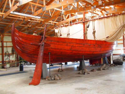 [Wooden ship with a large oar attached to the front and a low, wide mast with sail attached in the back. It is set on stands in a large, warehouse-like building.]